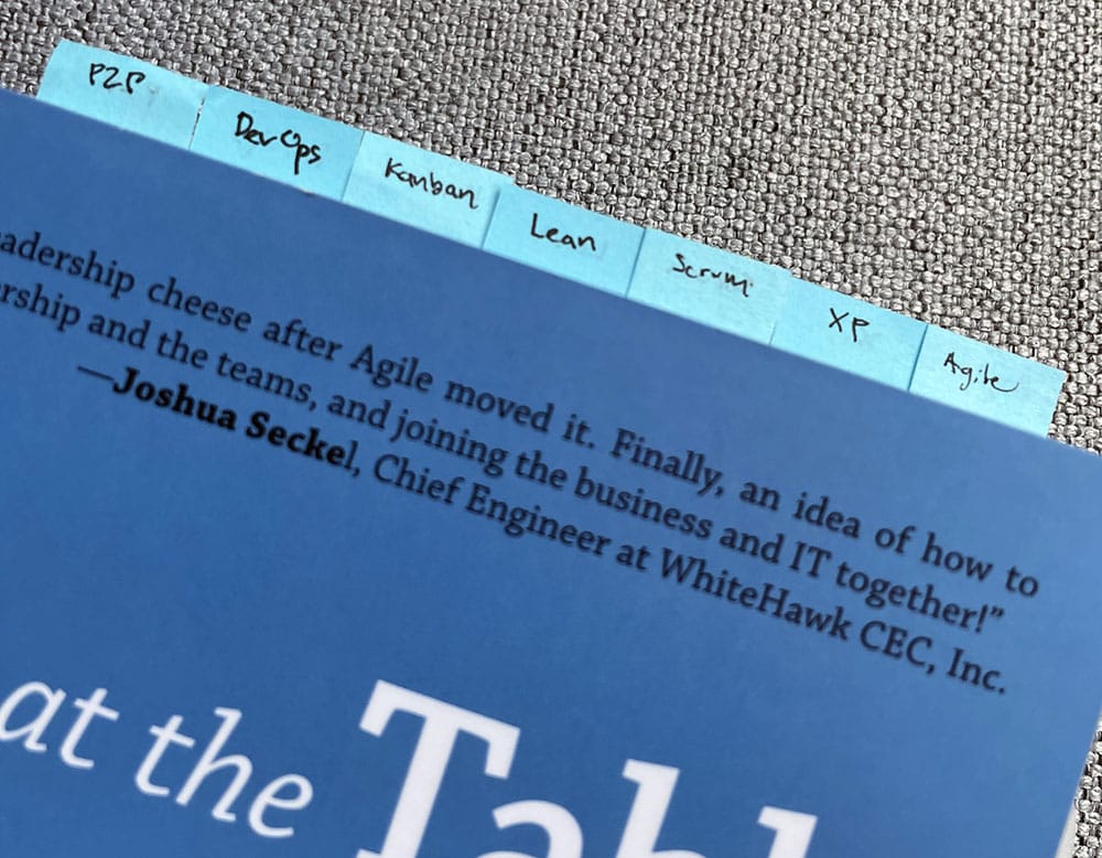 A photo of Lucy's copy of A Seat at the Table: IT Leadership in the Age of Agility, by Mark Schwartz. Only the upper part of the book is visible, and there are seven little blue tabs sticking out of the top of the book, labeled as follows: P2P, DevOps, Kanban, Lean, Scrum, XP, and Agile.